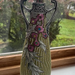 May include: A tall, olive green vase with two ornate handles, featuring a textured surface with vertical dotted lines, silver-toned floral accents, and small embedded colorful gems in shades of pink, blue, and green along the neck and body.
