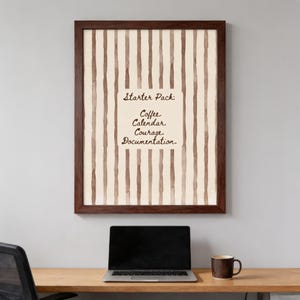 May include: A framed wall art print featuring vertical brown watercolour stripes on a cream background. Centered is a rectangular box with the text: Starter Pack: Coffee. Calendar. Courage. Documentation. The frame is dark brown wood.