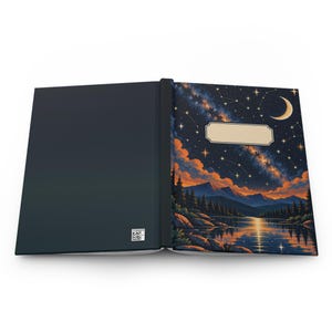 May include: An open hardcover notebook with a solid dark blue back cover and a front cover featuring a night landscape with a crescent moon, starry sky, mountains, and a lake. A blank rectangular label is centered on the front cover.