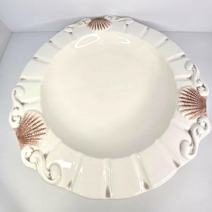Può includere: Una ciotola da portata rotonda in ceramica color bianco sporco con un bordo largo e scanalato. Il bordo presenta tre motivi a conchiglia in rilievo nei toni del marrone, intervallati da decorazioni a volute. Il centro è liscio.