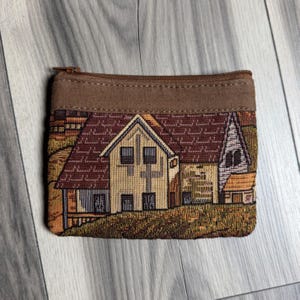 May include: A rectangular zippered pouch featuring a woven tapestry design of a house with a brown roof, beige walls, and dark windows. The top border of the pouch is made of solid brown fabric, and the overall scene depicts a building structure.