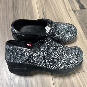 May include: A pair of black and white speckled clogs with a textured, stingray-like pattern. The shoes feature a black sole, a rounded toe, and a small Danish flag tag on the side. The interior is black with the brand name Sanita printed inside.