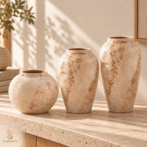 Pode incluir: Três vasos de cerâmica com acabamento texturizado e salpicado em tons de creme, bege e castanho ferrugem. O conjunto inclui um vaso arredondado de perfil baixo e dois vasos mais altos e alongados com aberturas estreitas, todos salpicados.