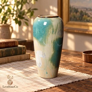 May include: A tall ceramic vase featuring a reactive glaze with blended shades of teal, cream, and white. The cylindrical vessel has a smooth, glossy finish with a dark brown rim and a tapered base, showcasing a fluid, painterly color transition.