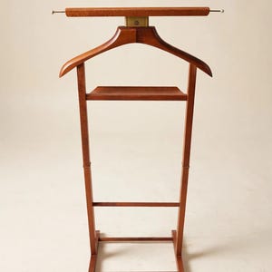 May include: A wooden valet stand featuring a contoured coat hanger at the top, a horizontal bar with small white end caps, and a flat shelf. The structure is supported by two vertical legs connected by a lower crossbar and a rectangular base.