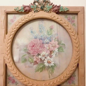 May include: A square wall frame with a circular center featuring a floral print of pink roses, white daisies, and blue flowers. The frame is light pink with carved leaf details and a decorative bow at the top. It measures approximately 10 inches tall by 9 inches wide.