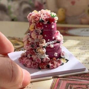 May include: A miniature two-tier cake in a deep magenta hue, decorated with an abundance of small, sculpted pink, cream, and red flowers, white pearl accents, and a single red strawberry on top, set on a square white base.