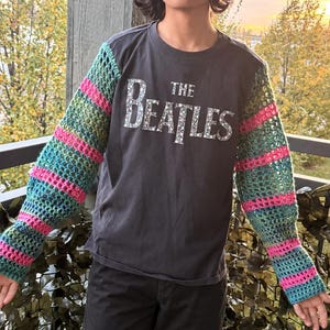 May include: A long-sleeve shirt featuring a dark gray torso with the text THE BEATLES printed in white. The sleeves are made of a crocheted mesh fabric with horizontal stripes in alternating colors of teal, green, and bright pink.