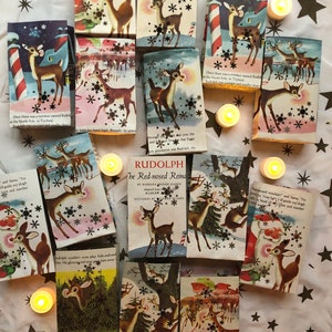 May include: A collection of vintage Rudolph the Red-Nosed Reindeer books with illustrations of Rudolph and Santa Claus. The books are arranged on a white fabric background with gold stars and small white tea lights.