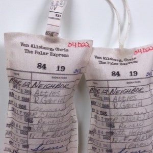 May include: Two white fabric book card pockets with blue handwritten text. The text includes the title "The Polar Express" and the date "Mar 12 Neighbor".