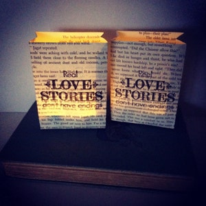 May include: Two small paper bags with a warm glow from candles inside. The bags are decorated with text from a book, including the words "Real Love Stories" and "don't have endings".