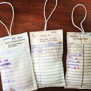 May include: Three vintage library checkout cards with white paper and blue lines. The cards are tied with white string and have handwritten information. The top card reads "CHAWTON 80 966 Moore, Clement Clarke The Night Before Christmas". The middle card reads "Seuss, Dr. How the Grinch Stole Christmas 04712". The bottom card reads "DICKENS A Christmas Carol MONTCLAIR 150 D7062 7669 6591 MR 10415 MR JAN 2 1981 PATION, JOHN FREDERICK JAN 16 1981 N."