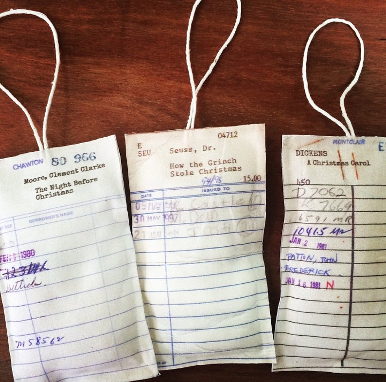May include: Three vintage library checkout cards with white paper and blue lines. The cards are tied with white string and have handwritten information. The top card reads "CHAWTON 80 966 Moore, Clement Clarke The Night Before Christmas". The middle card reads "Seuss, Dr. How the Grinch Stole Christmas 04712". The bottom card reads "DICKENS A Christmas Carol MONTCLAIR 150 D7062 7669 6591 MR 10415 MR JAN 2 1981 PATION, JOHN FREDERICK JAN 16 1981 N."