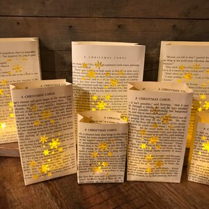 May include: A collection of handmade paper luminaries crafted from pages of the book "A Christmas Carol." Each luminary is adorned with gold glitter snowflake accents. The text "A Christmas Carol" is visible on the pages.