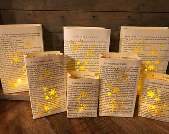 May include: A collection of handmade paper luminaries crafted from pages of the book "A Christmas Carol." Each luminary is adorned with gold glitter snowflake accents. The text "A Christmas Carol" is visible on the pages.