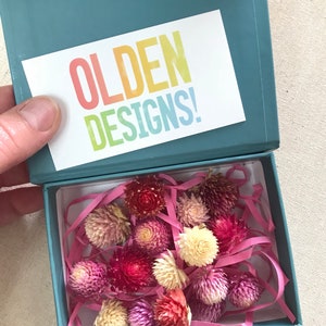 May include: A teal gift box with a white card that says "OLDEN DESIGNS!" inside the box are dried flowers in shades of pink, red, and white. The flowers are surrounded by pink tissue paper.