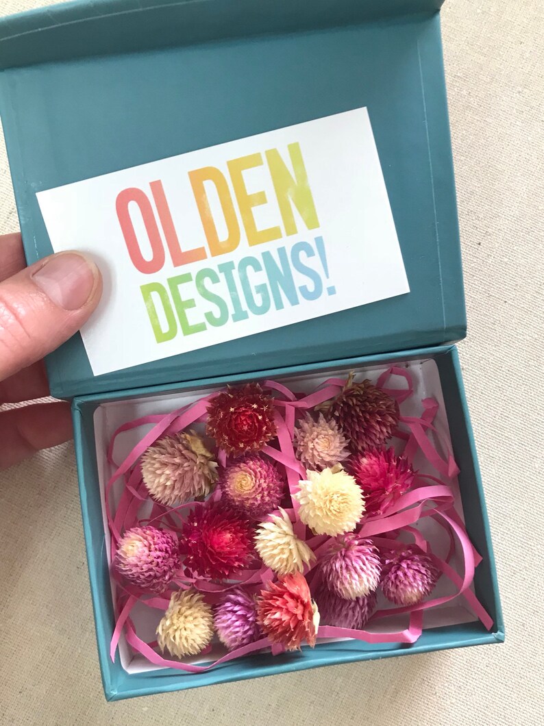 May include: A teal gift box with a white card that says "OLDEN DESIGNS!" inside the box are dried flowers in shades of pink, red, and white. The flowers are surrounded by pink tissue paper.