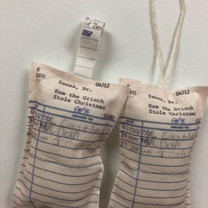 May include: Two white fabric ornaments shaped like library card pockets with the text "Seuss, Dr. How the Grinch Stole Christmas 94/95 Issued to 15.00" printed on them. The ornaments are hanging from a white string.
