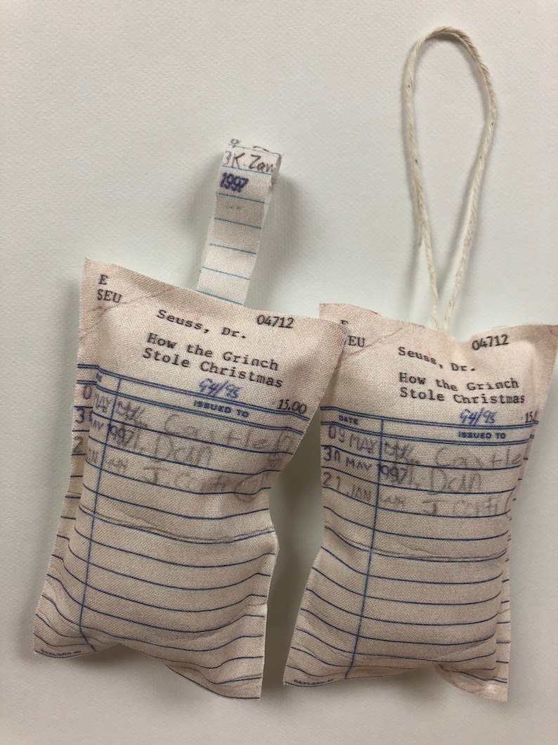May include: Two white fabric ornaments shaped like library card pockets with the text "Seuss, Dr. How the Grinch Stole Christmas 94/95 Issued to 15.00" printed on them. The ornaments are hanging from a white string.