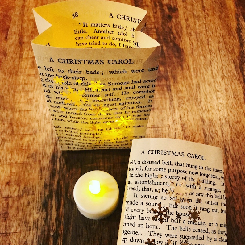 May include: A close-up of a book titled "A Christmas Carol" with a small, lit candle in the foreground. The book is open to a page with text about Scrooge and his transformation. The candle is on a wooden surface.