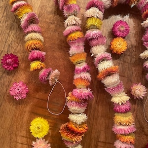 May include: Three strands of dried flower garland made with pink, white, orange, and yellow flowers. The garlands are arranged in a circle on a wooden surface.