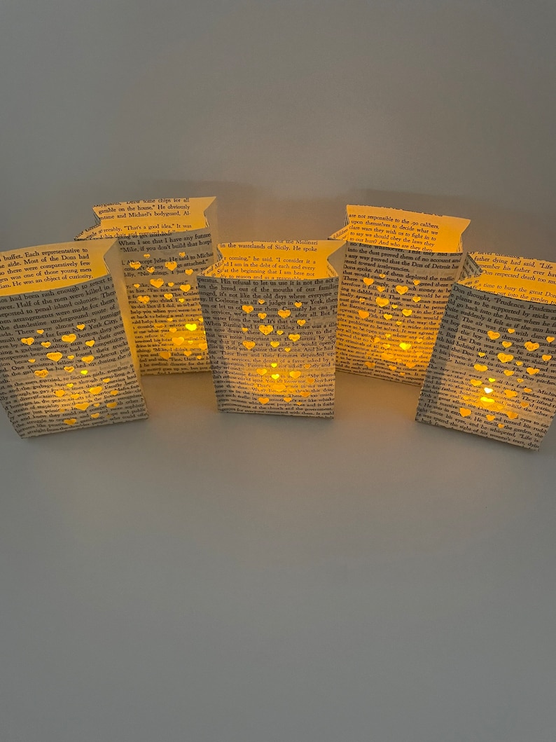 May include: Six paper lanterns with a warm glow, each made from a book page with cut-out hearts. The lanterns are strung together and hanging from a ceiling.