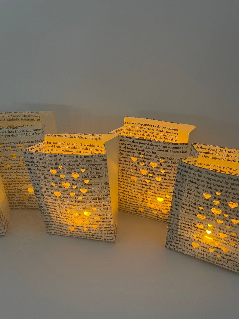 May include: A collection of small paper bags with cut-out hearts and a warm glow emanating from inside. The bags are filled with text from a book, and the text is visible through the cut-out hearts. The bags are arranged in a row on a white surface.