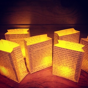 May include: A collection of small, paper lanterns with a warm, yellow glow. Each lantern is made from a recycled book page, with text visible on the outside. The lanterns are arranged on a wooden surface.