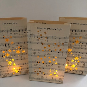 May include: Three paper lanterns with a Christmas theme. Each lantern features a different carol sheet music with gold stars and snowflakes. The lanterns are lit from within, creating a warm glow.