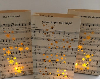 May include: Three paper lanterns with a Christmas theme. Each lantern features a different carol sheet music with gold stars and snowflakes. The lanterns are lit from within, creating a warm glow.
