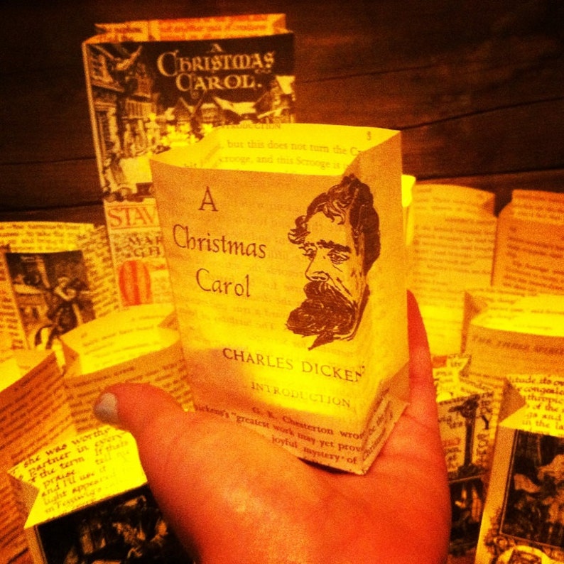 May include: A close-up of a hand holding a yellow paper bag with the title "A Christmas Carol" by Charles Dickens. The bag is decorated with a black and white illustration of Charles Dickens.