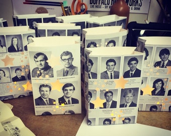Puede incluir: Una colección de bolsas de papel blancas decoradas con fotos en blanco y negro de personas y estrellas doradas. Las bolsas están dispuestas en filas sobre una superficie de madera. El texto "BUFFALO RI WORKS" es visible en un cartel en el fondo.