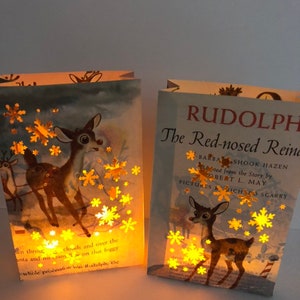May include: Two illuminated book page lanterns featuring deer illustrations and snowflake cutouts. One lantern displays the text "RUDOLPH The Red-nosed Reindeer". The warm glow highlights the vintage book pages, creating a cozy, festive ambiance.