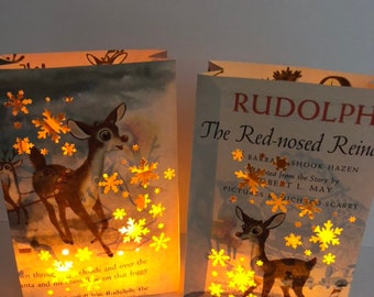May include: Two illuminated book page lanterns featuring deer illustrations and snowflake cutouts. One lantern displays the text "RUDOLPH The Red-nosed Reindeer". The warm glow highlights the vintage book pages, creating a cozy, festive ambiance.