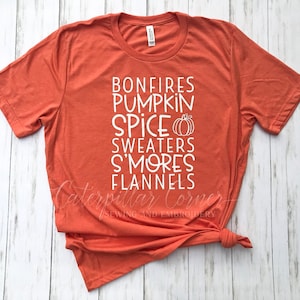 May include: A burnt orange t-shirt with white text that reads "Bonfires Pumpkin Spice Sweaters S'mores Flannels". A small pumpkin graphic is next to the word "Spice".