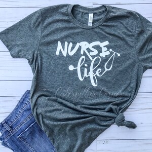 Op de afbeelding: Een heidegrijs T-shirt met de tekst "NURSE life" in witte letters. De tekst is in een gestileerd lettertype en bevat een stethoscoopgrafiek.