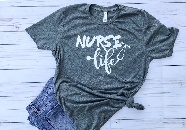 Op de afbeelding: Een heidegrijs T-shirt met de tekst "NURSE life" in witte letters. De tekst is in een gestileerd lettertype en bevat een stethoscoopgrafiek.