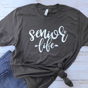 May include: A dark gray t-shirt with the text "senior life" in white lettering with an arrow on either side of the word "life". The shirt is tied in a knot at the bottom.