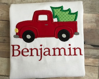May include: A white t-shirt with a red truck carrying a green Christmas tree and the name "Benjamin" embroidered in red.