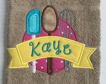 Può includere: Uno stendardo ricamato giallo con il nome "Kaye" su un asciugamano marrone. Lo stendardo è sopra un cerchio rosa con pois bianchi e tre utensili da cucina ricamati: una spatola blu, un cucchiaio di legno marrone e una frusta argentata.