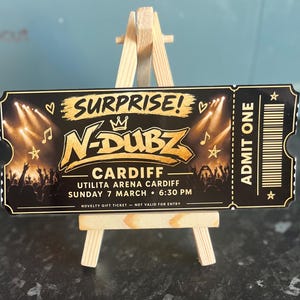 May include: A black novelty gift ticket for an N-Dubz concert at Utilita Arena Cardiff on Sunday 7 March at 6:30 PM. The ticket features gold text, a crown icon, stars, musical notes, and a concert crowd graphic, displayed on a small wooden easel.