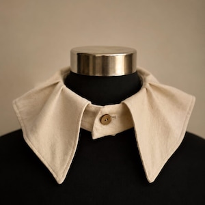 May include: A detachable collar in a light beige, off-white fabric featuring a pointed, wide-spread design with soft pleats at the neckline. The collar is secured at the front center with a single, small, round, brown button and a buttonhole.