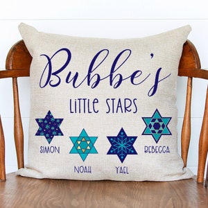May include: A beige linen pillow with the text "Bubbe's Little Stars" in navy blue. Four blue and teal Star of David designs are arranged across the pillow, each with a name underneath: Simon, Noah, Yael, and Rebecca.