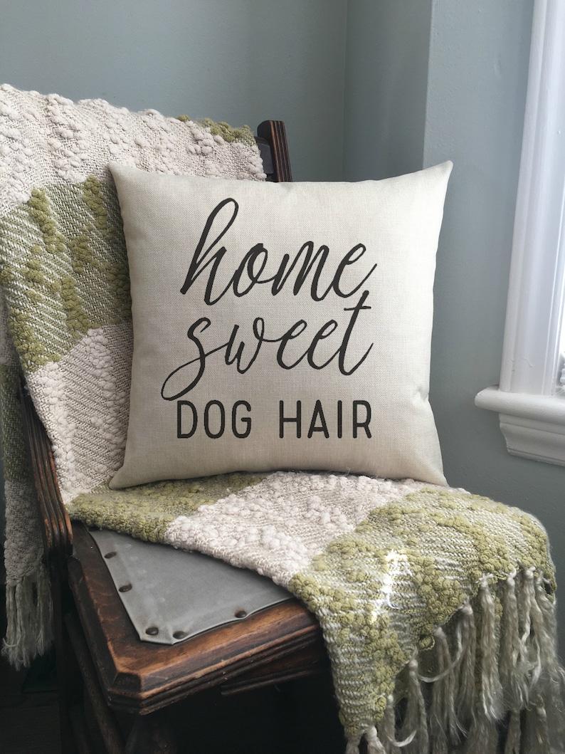 May include: A beige throw pillow with the text "home sweet dog hair" in black lettering. The pillow is sitting on a wooden chair with a green and beige throw blanket.