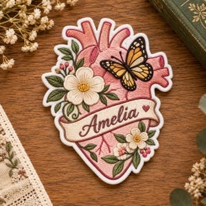 May include: An embroidered patch shaped like an anatomical heart in shades of pink. It features a yellow and black butterfly, white flowers with green leaves, and a cream-colored banner across the center with the name Amelia in brown.