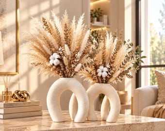May include: Two off-white, arch-shaped ceramic vases holding arrangements of dried pampas grass, fluffy white cotton bolls, and eucalyptus stems. The vases have a smooth, matte finish and a distinct U-shape, creating a balanced display.