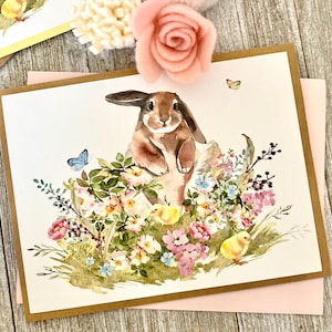 May include: Watercolor Easter greeting card featuring a brown bunny surrounded by colorful flowers, butterflies, and two yellow chicks. The card has a brown border and is on a pink envelope.