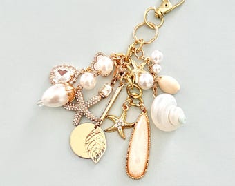 May include: A gold-toned keychain with various charms, including pearls, seashells, and starfish. The charms are attached to a gold chain and clasp. The keychain also features a gold leaf and a teardrop-shaped charm.