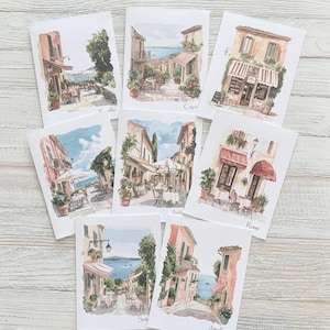 May include: A collection of watercolor greeting cards featuring Italian coastal scenes. Each card depicts a different town, including Capri, Amalfi, and Rome, with cafes and buildings in shades of pink, orange, and blue. The cards are arranged on a light wood surface.