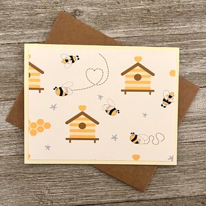 Puede incluir: Una tarjeta amarilla con fondo blanco y un patr&oacute;n de colmenas, abejas y corazones. La tarjeta tiene un sobre marr&oacute;n claro.
